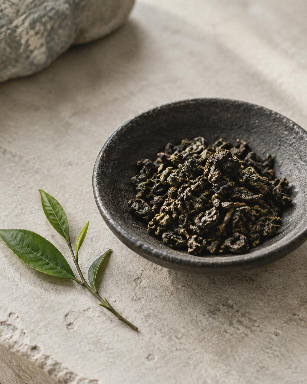 Loose rolled tea leaves in a dark bowl beside fresh green leaves on stone.